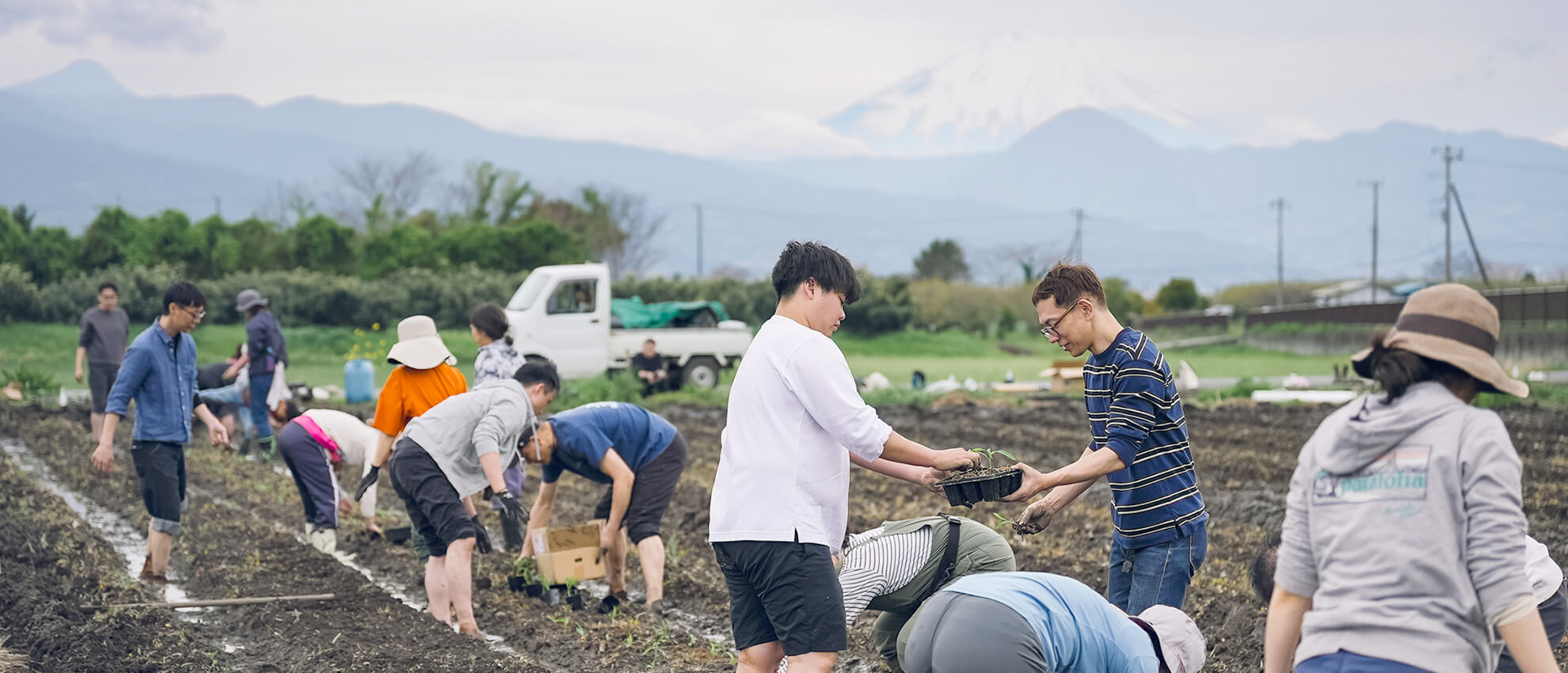 仲間とともに畑で植え付け作業をする人々。雪をいただく山々を背景に、自然と協力しながら大地に向き合う姿。